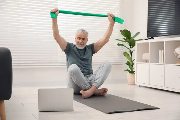 Senior man doing exercise with fitness elastic band near laptop on mat at home