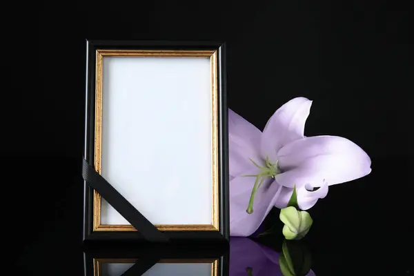 Funeral photo frame with ribbon and violet lily on table against black background