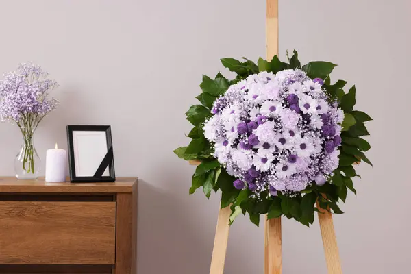 Wreath of violet and white flowers, photo frame with black ribbon in room. Funeral attributes