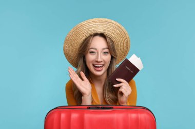 Açık mavi arka planda pasaportu, bileti ve bavulu olan mutlu genç bir kadın.