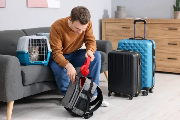 Travel with pet. Man putting headphones into backpack near carrier with cute cat at home