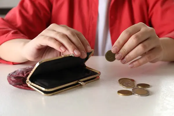 Poor woman putting coin into empty wallet at white table, closeup