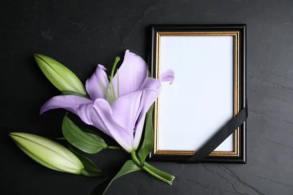 Funeral photo frame with ribbon and violet lily on black table, flat lay