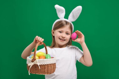 Paskalya kutlaması. Tavşan kulaklı şirin kız yeşil arka planda boyalı yumurta sepeti tutuyor.