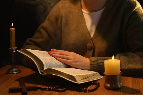 Woman reading Bible at table with burning candles, closeup