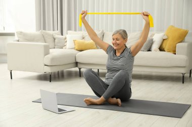 Senior woman doing exercise with fitness elastic band near laptop on mat at home clipart