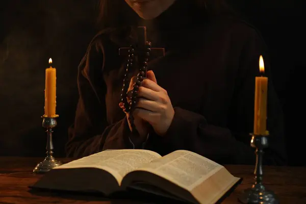 Woman praying at table with burning candles and Bible, closeup