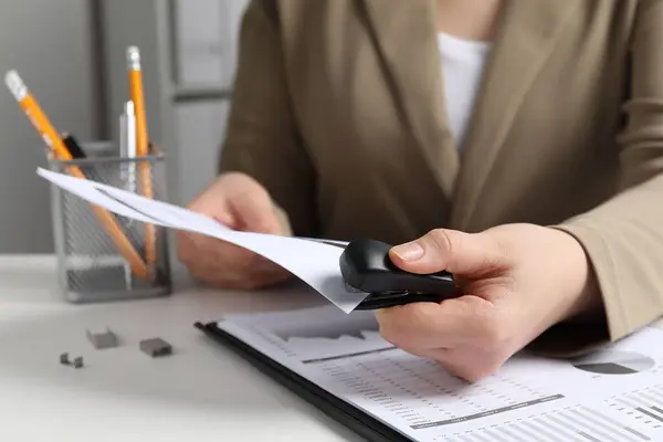 Woman with papers using stapler at white table indoors, closeup