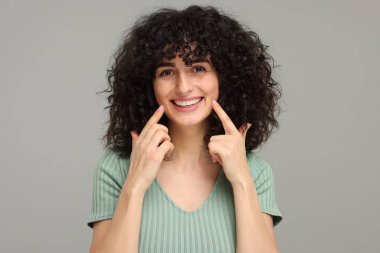 Gri arka planda beyazlatıcı şeritle dişlerini gösteren genç bir kadın.