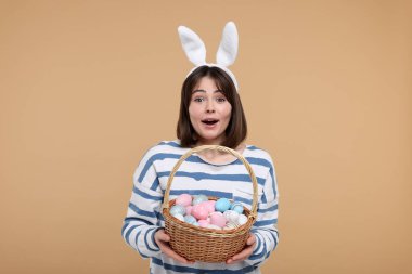 Paskalya kutlaması. Tavşan kulaklı ve bej arka planda boyanmış yumurta dolu sepeti olan şaşırmış bir kadın.