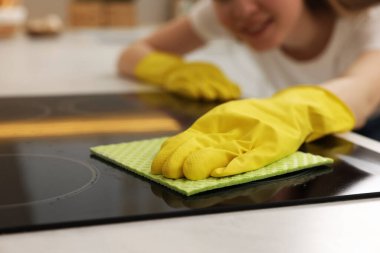 Elinde sprey şişesi ve mikrofiber bez olan bir kadın mutfakta elektrikli soba temizliyor.