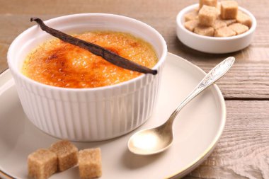 Lezzetli creme brulee kasede, vanilya kapsülü, kaşık ve şeker küpleri ahşap masada, yakın plan