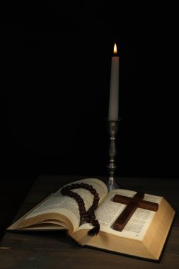 Kilise mumu, İncil, tespih boncukları ve ahşap masada siyah arka plana karşı haç.