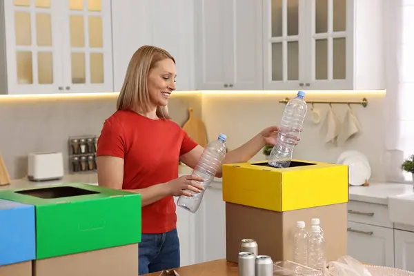 Garbage sorting. Smiling woman throwing plastic bottle into cardboard box in kitchen