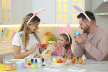 Paskalya kutlaması. Mutfaktaki beyaz mermer masada yumurta boyayan tavşan kulaklı mutlu aile.