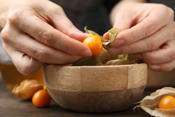Woman peeling physalis fruit from calyxes at wooden table, closeup