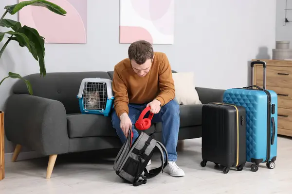 Travel with pet. Man putting headphones into backpack near carrier with cute cat at home