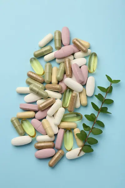 Different vitamin pills and green branch on light blue background, flat lay