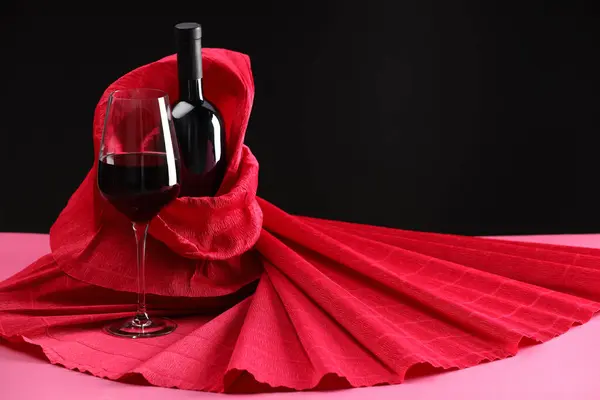Stylish presentation of delicious red wine in bottle and glass on pink table against black background. Space for text