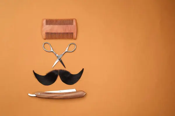 Artificial moustache and barber tools on orange background, flat lay. Space for text