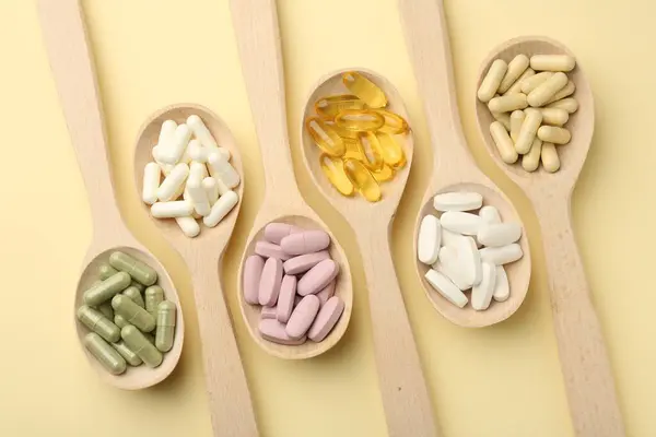 Different vitamin pills in spoons on pale yellow background, flat lay