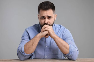 Açık gri arka planda, ahşap masada oturan üzgün adamın portresi.