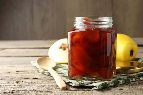Tasty homemade quince jam in jar, spoon and fruits on wooden table, closeup. Space for text