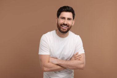 Çapraz kollu yakışıklı adam açık kahverengi arka planda gülüyor.