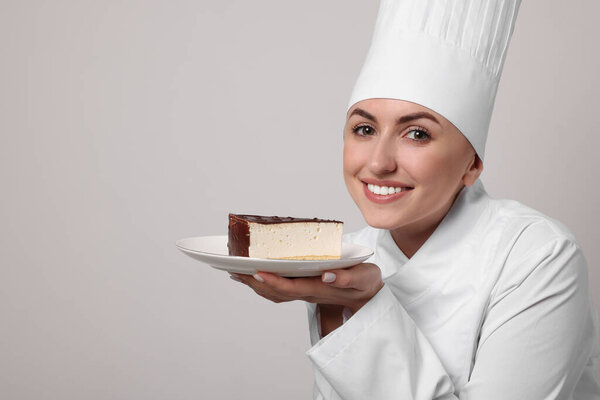 Happy professional confectioner in uniform holding delicious cheesecake on light grey background. Space for text
