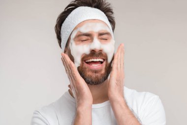 Saçbandı olan bir adam açık gri arka planda yüzünü yıkıyor.