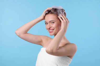Saçı açık mavi arka planda şampuanla yıkanan mutlu genç bir kadın.