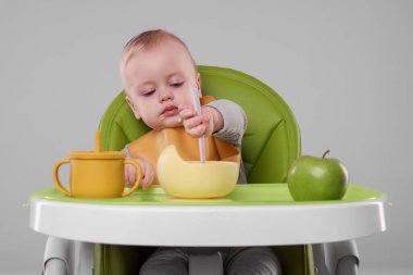 Gri arka planda, yüksek sandalyede sağlıklı yiyecekler yiyen şirin bir bebek.