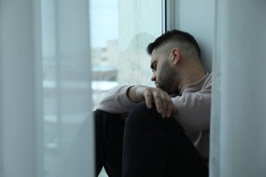 Evdeki pencerenin yanında oturan üzgün adam.