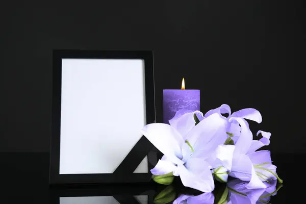 Funeral photo frame with ribbon, violet lily flowers and candle on dark table against black background