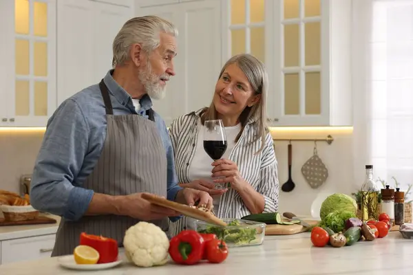 Happy senior couple cooking together in kitchen. Woman with glass of wine near her husband