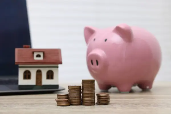 House model, piggy bank and stacked coins on light wooden table, selective focus