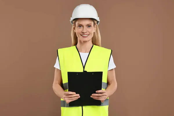 Engineer in hard hat holding clipboard on brown background