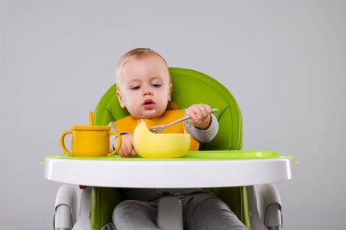 Gri arka planda, yüksek sandalyede sağlıklı yiyecekler yiyen şirin bir bebek.