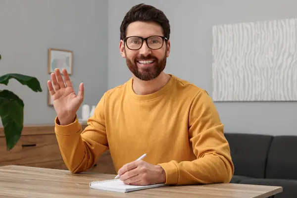 Man in glasses greeting someone at wooden table indoors