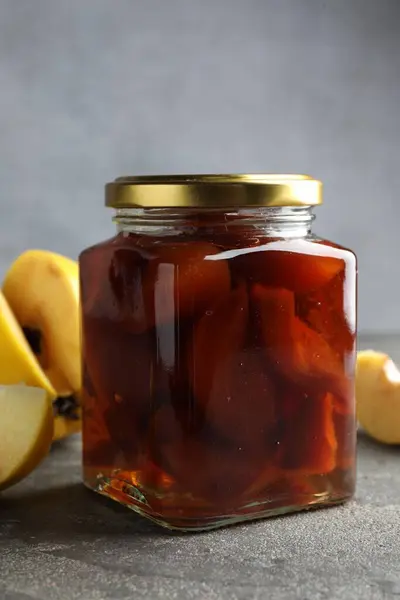 Tasty homemade quince jam in jar and fruits on grey textured table, closeup