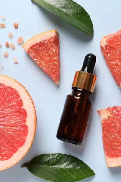 Grapefruit essential oil in bottle, sea salt, leaves and cut fruit on light blue table, flat lay