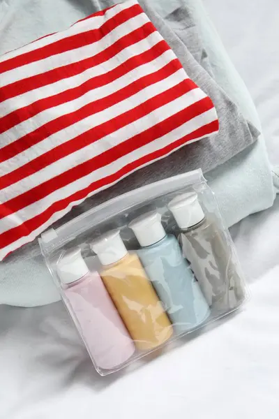 Cosmetic travel kit. Plastic bag with small containers of personal care products and stack of clothes on bed