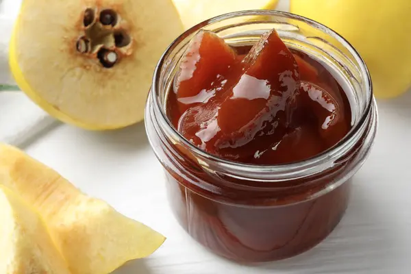 Tasty homemade quince jam in jar and fruits on white wooden table, closeup