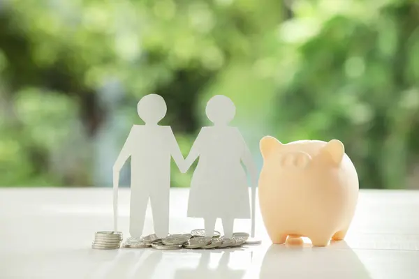 Pension savings. Figure of senior couple, piggy bank and coins on white table against blurred green background