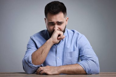 Açık gri arka planda, ahşap masada oturan üzgün adamın portresi.