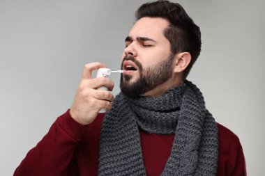 Gri arka planda boğaz spreyi kullanan atkılı genç bir adam.