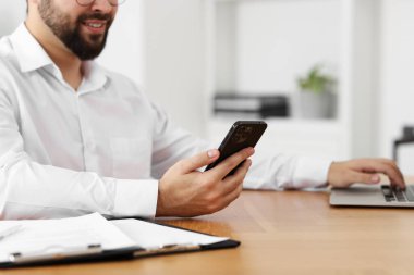 Ofisteki ahşap masada dizüstü bilgisayarla çalışırken akıllı telefon kullanan genç adam.