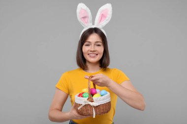 Paskalya kutlaması. Tavşan kulaklı mutlu kadın ve gri arka planda boyanmış yumurta dolu hasır sepet.