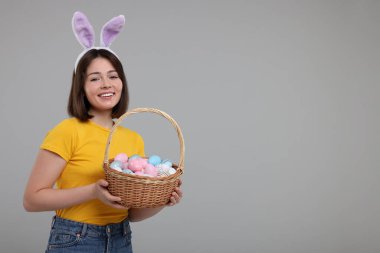 Paskalya kutlaması. Tavşan kulaklı mutlu kadın ve gri arka planda boyanmış yumurta dolu hasır sepet, mesaj için yer