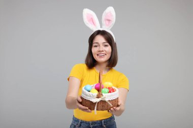 Paskalya kutlaması. Tavşan kulaklı mutlu kadın ve gri arka planda boyanmış yumurta dolu hasır sepet.